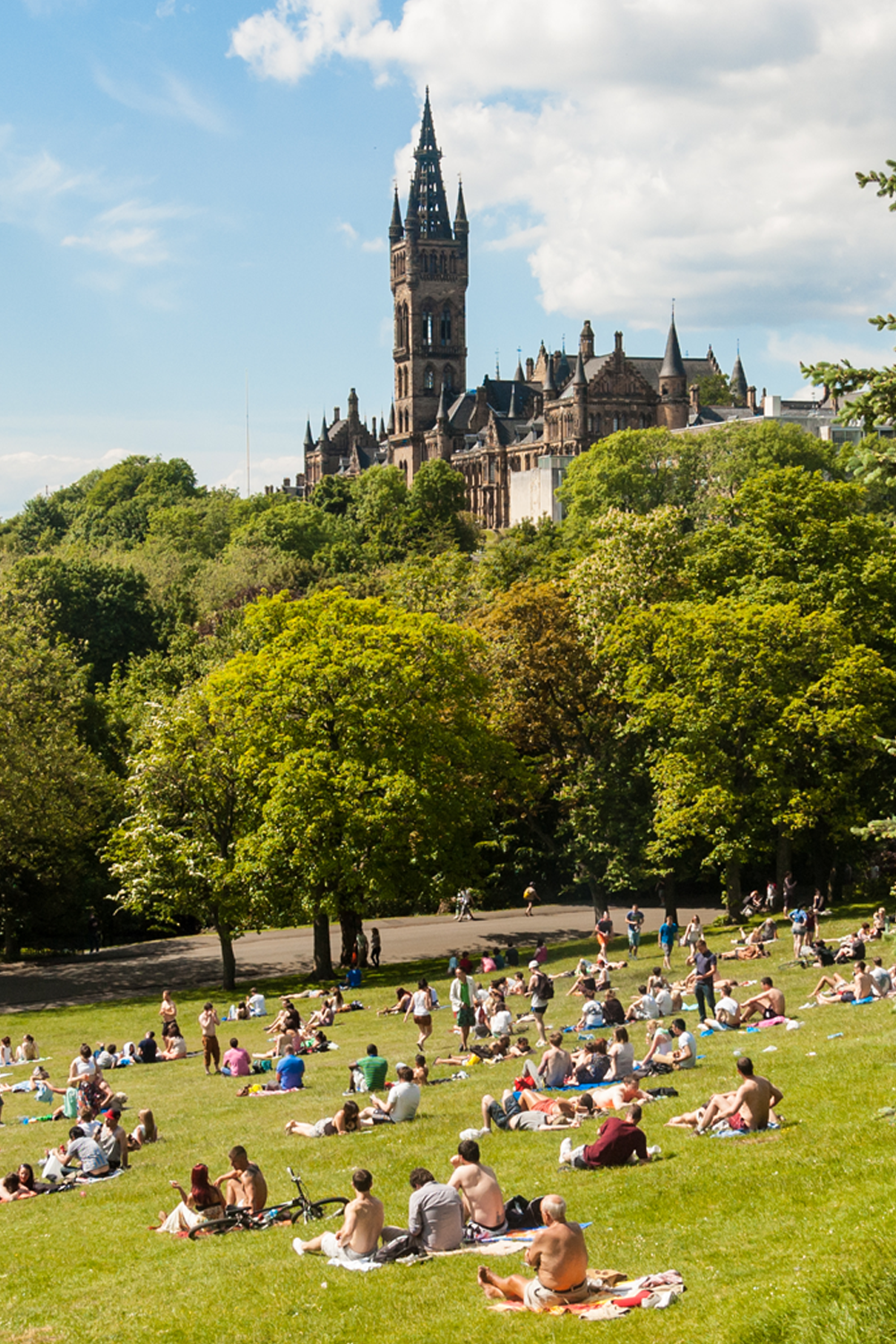 View of Kelvingrove Park full of people enjoying the Scottish summer with the main building of Glasgow University on the top of the hill.