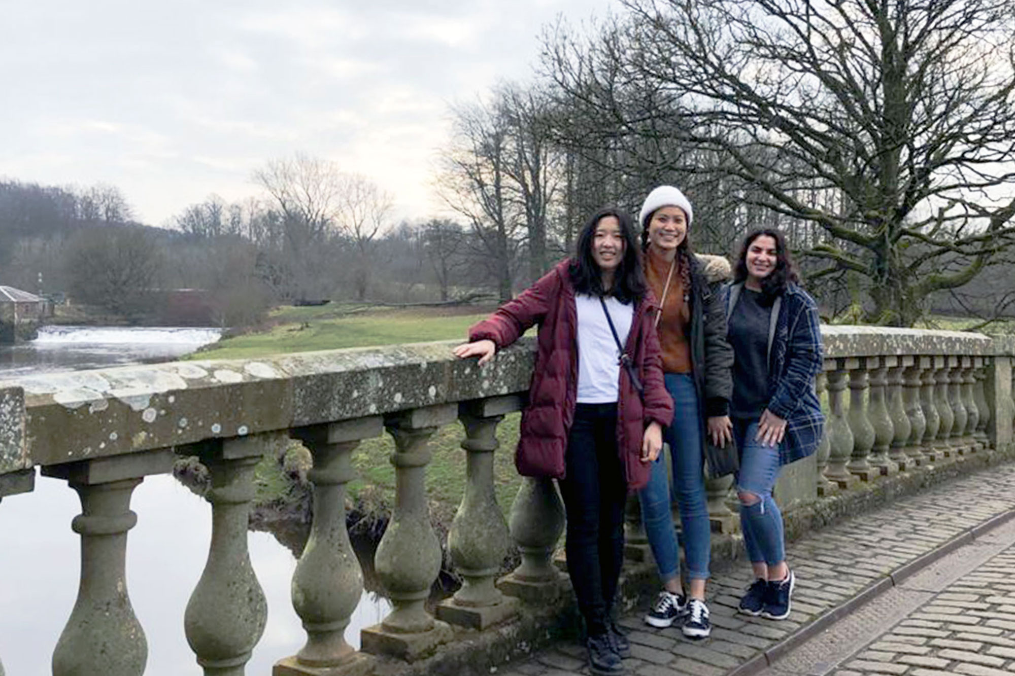 Christine Choy smiling at the camera with two friiends standing on a bridge over a river in parkland, Glasgow