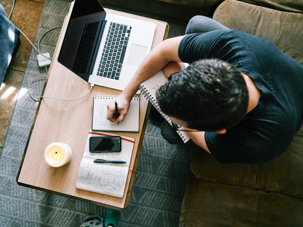 Student sitting on a couch and taking notes next to his computer