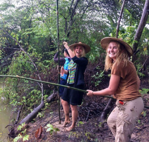 Two students laughing and smiling for camera as stand on a riverbank with bamboo fishing poles.