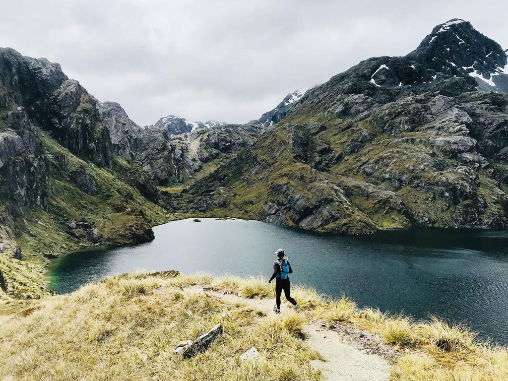 student running down a pathway near a lake and lush green mountains in New Zealand.