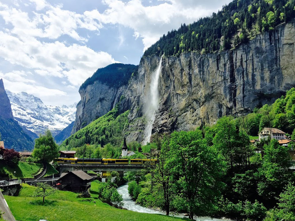 Nature view of a Swiss farm town with a train crossing a river near a mountain.