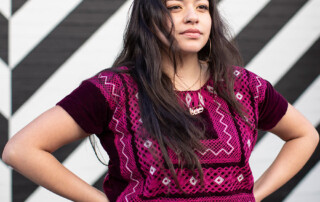 Close up of young Latina wearing magenta ethnic top, standing with hands on hips.