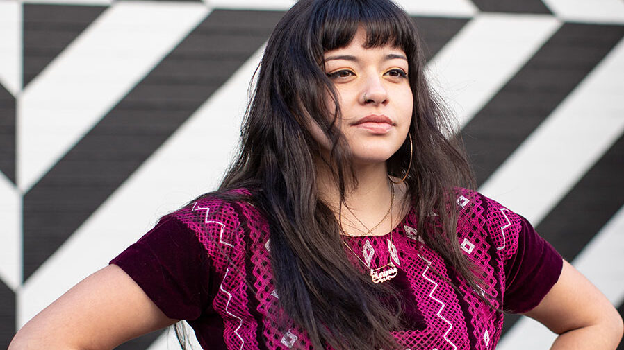 Close up of young Latina wearing magenta ethnic top, standing with hands on hips.