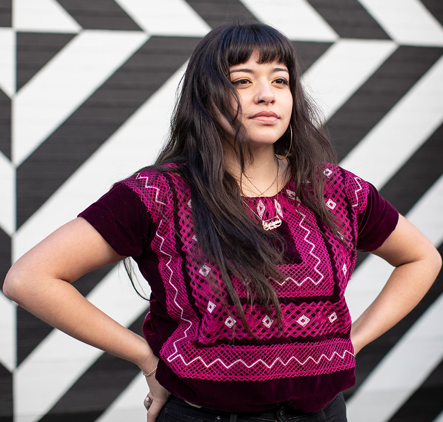 Close up of young Latina wearing magenta ethnic top, standing with hands on hips.