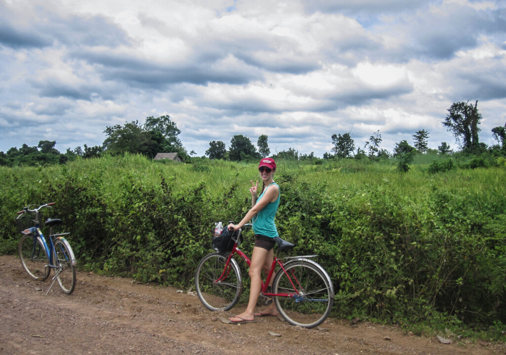 Kristin poses for a photo while riding a bike on a dirt road alongside a lush green field.