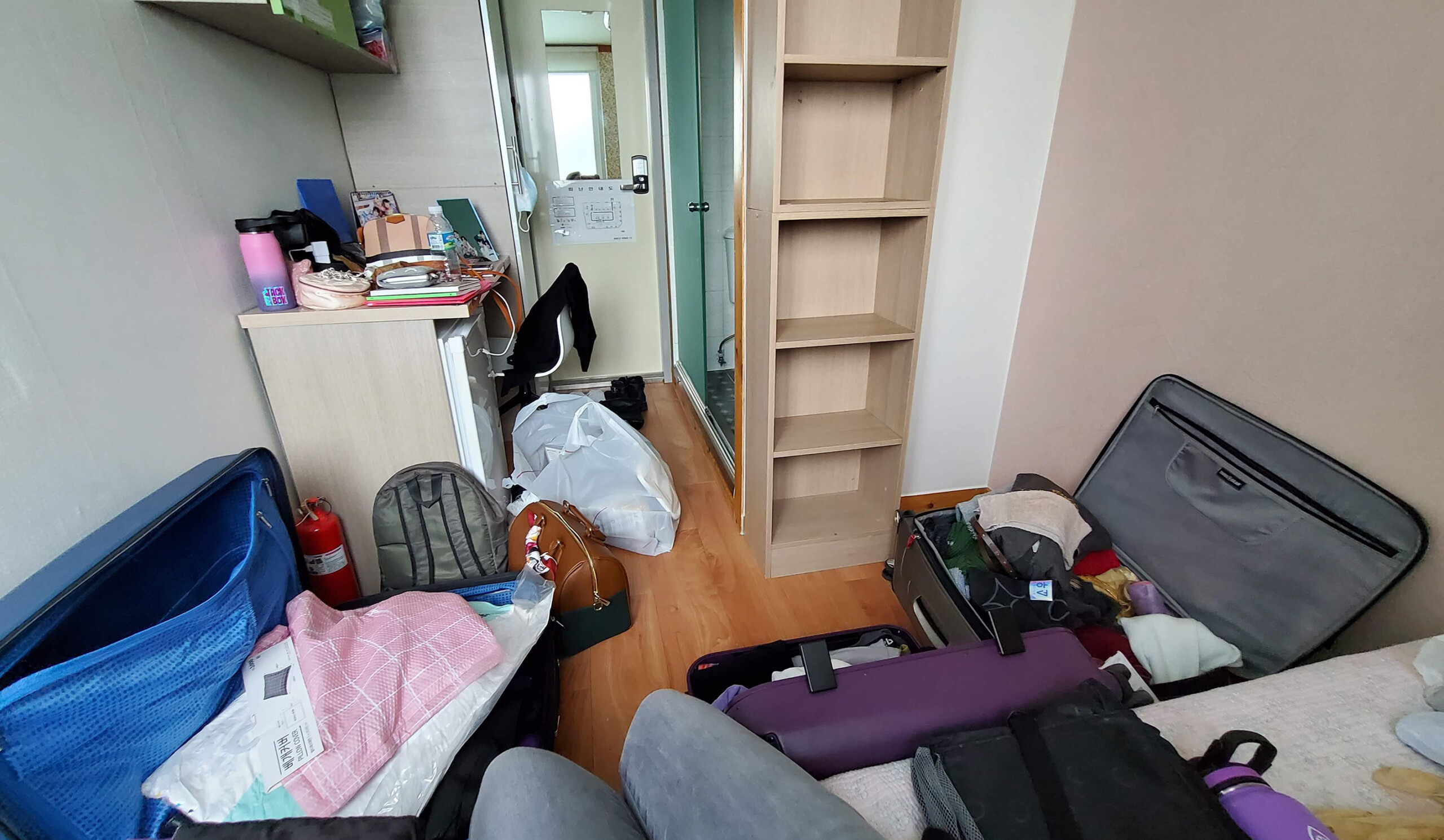 A picture of a goshiwon room with a twin sized bed, a large window, a private bathroom, cabinets, a closet, desk, mini fridge, and shelves.