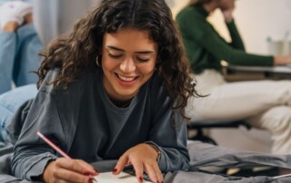 A female presenting teen lies on a bed and writes on a notepad in a dorm with a friend studying in the background.