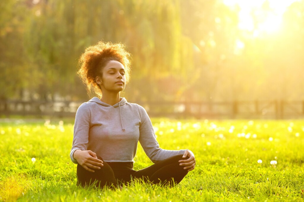 Female-presenting person sits in a grassy area and meditates with legs crossed.