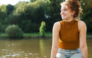 Young female presenting person sits on a wall with a lake behind them and smiles on a sunny day in London.