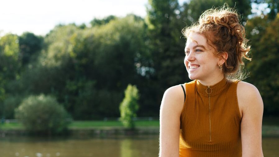 Young female presenting person sits on a wall with a lake behind them and smiles on a sunny day in London.