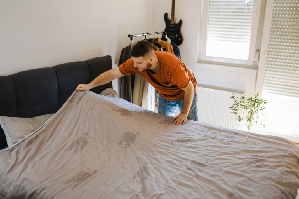 A male presenting person places a sheet over their bed.