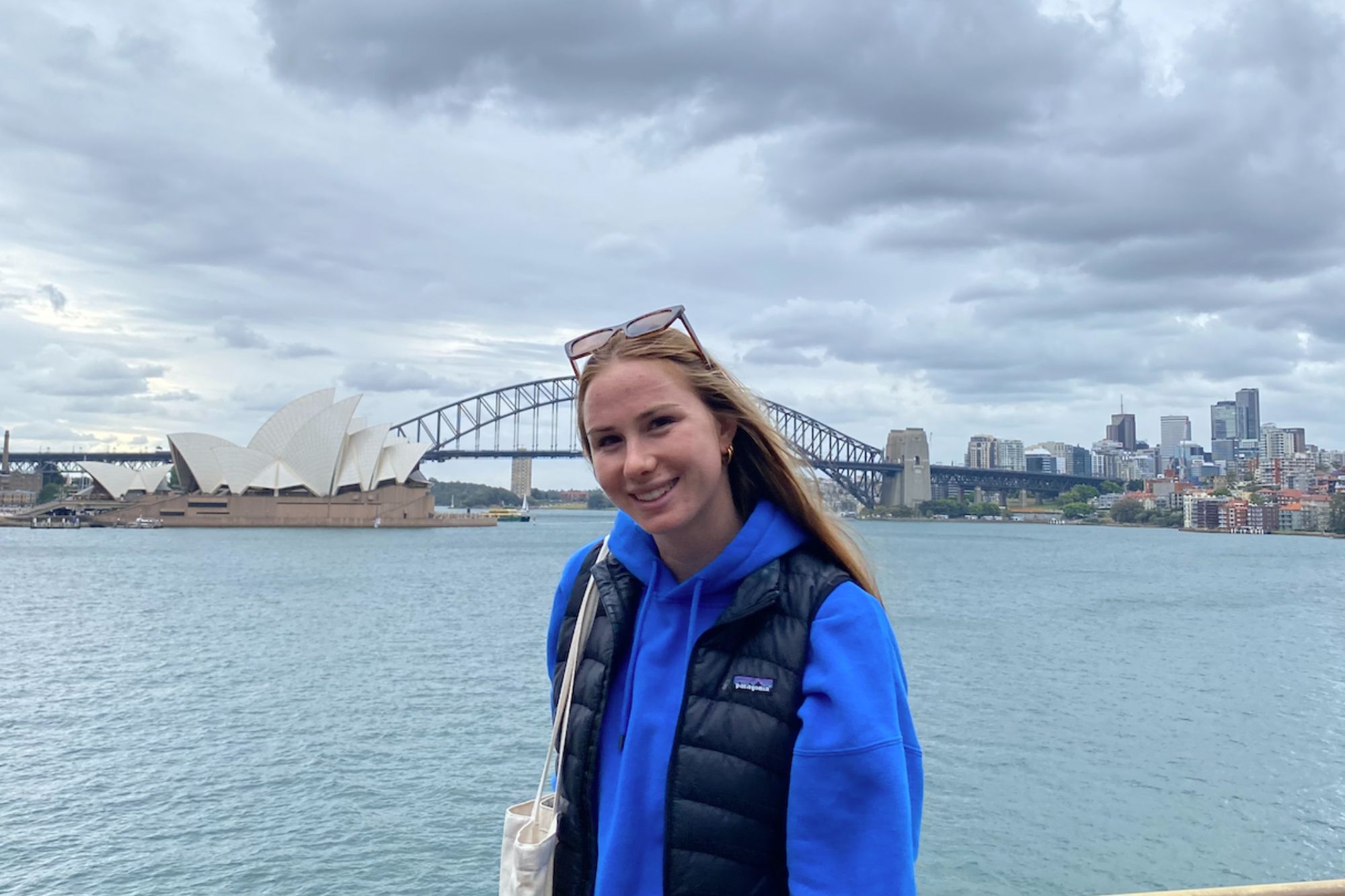 A student smiles in front of the Sydney Skyline, including the Symphony Orchestra behind them.