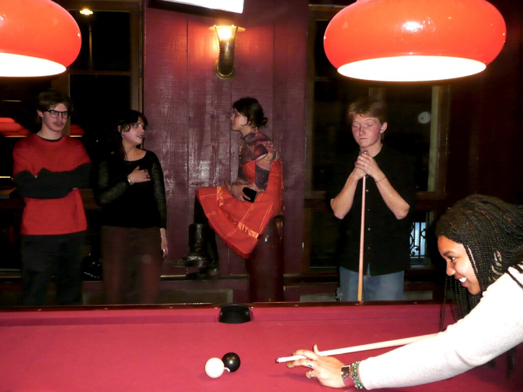 Person playing table-top pool on a red pool table and students looking on in the background