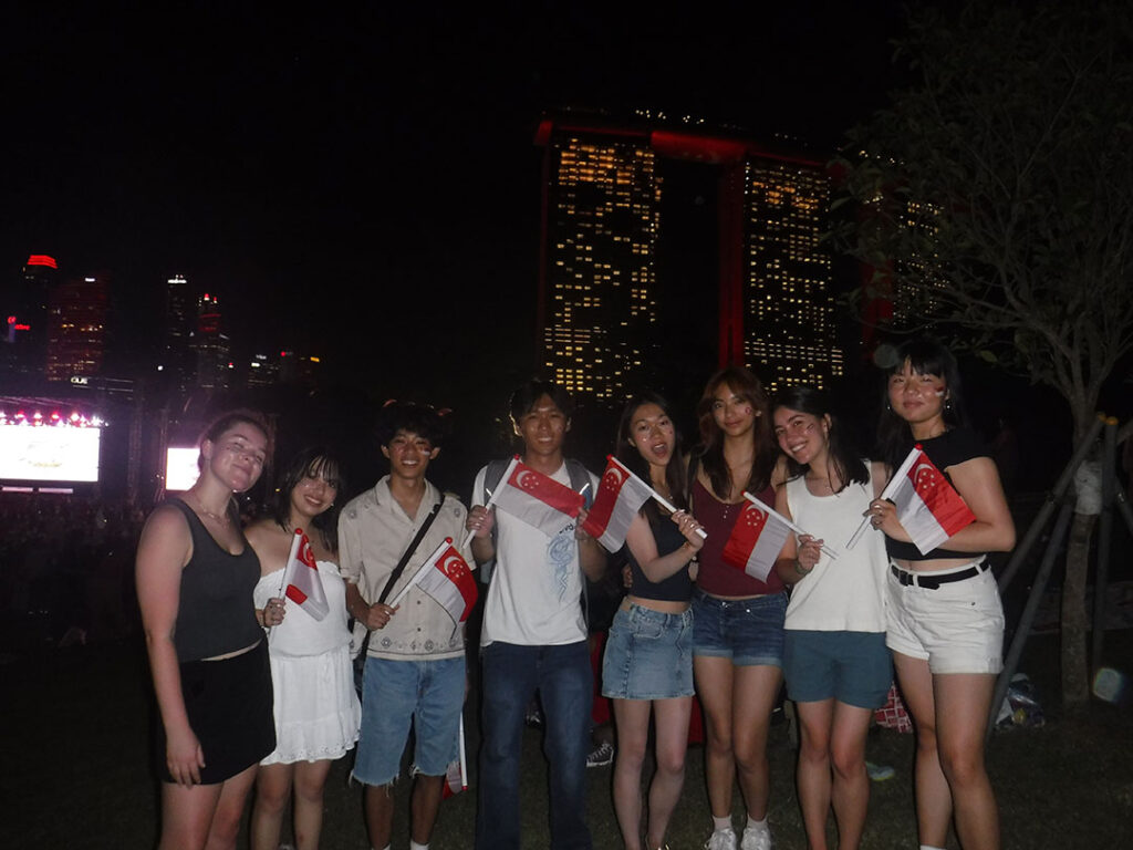 A group of eight young people stands together at night, smilihng and holding Singapore flags. The illuminated Marina Bay Sands is visible in the background.