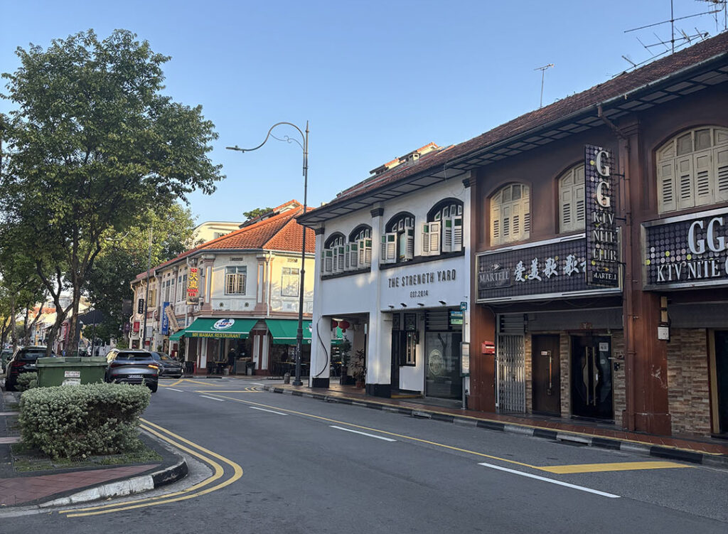 Street view of colorful shophouses with signage under a clear blue sky. A quiet road with a few parked cars and greenery lining the sidewalk.