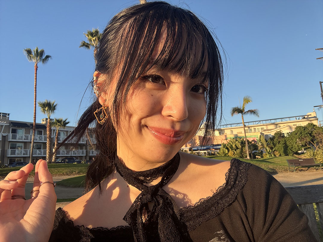Smiling woman with dark hair and bangs poses outdoors in sunlight, wearing a black top and gold earrings. Palm trees and buildings in the background.