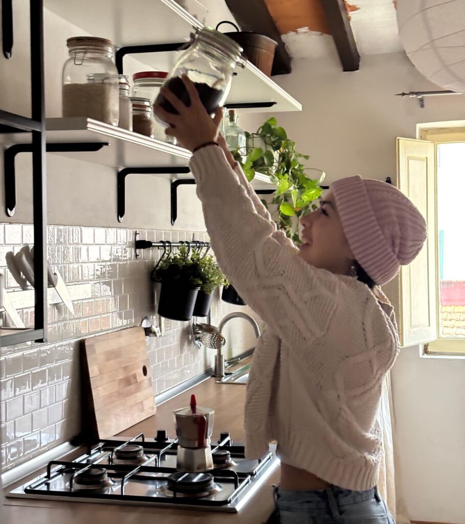 student in pink beanie hat reaching up to high shelf to grab coffee grounds while her moka pot is on the stove next to her